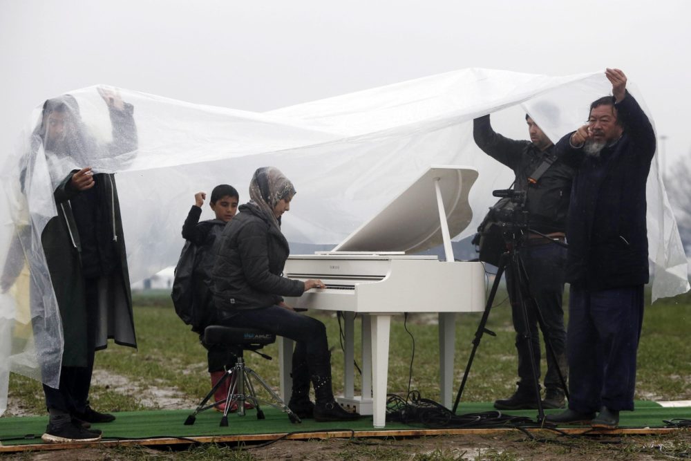 epa05208051 Chinese contemporary artist, Ai Weiwei (R) protects himself and others from the rain, as Nour Al Khizam (C) from the city Deirez Zor, Syria, plays the piano during a performing at a refugee camp, at the border of Greece and the Former Yugoslav Republic of Macedonia (FYROM), near Idomeni, Northern Greece, 12 March 2016. Greece has some 36,000 migrants trapped due to entry restrictions imposed by Macedonia in recent months. EPA/YANNIS KOLESIDIS